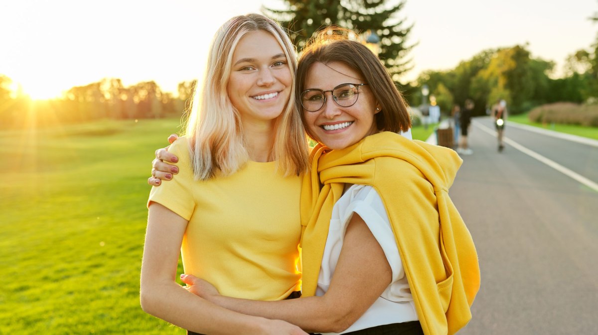 Ältere Mutter und Tochter im Teenageralter posieren bei Sonnenuntergang vor einer Wiese.