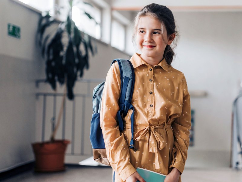 Mädchen, ca. 11 Jahre alt, steht in einem Schulgebäude, den Rucksack über einer Schulter gelegt, in den Händen ein Buch haltend.