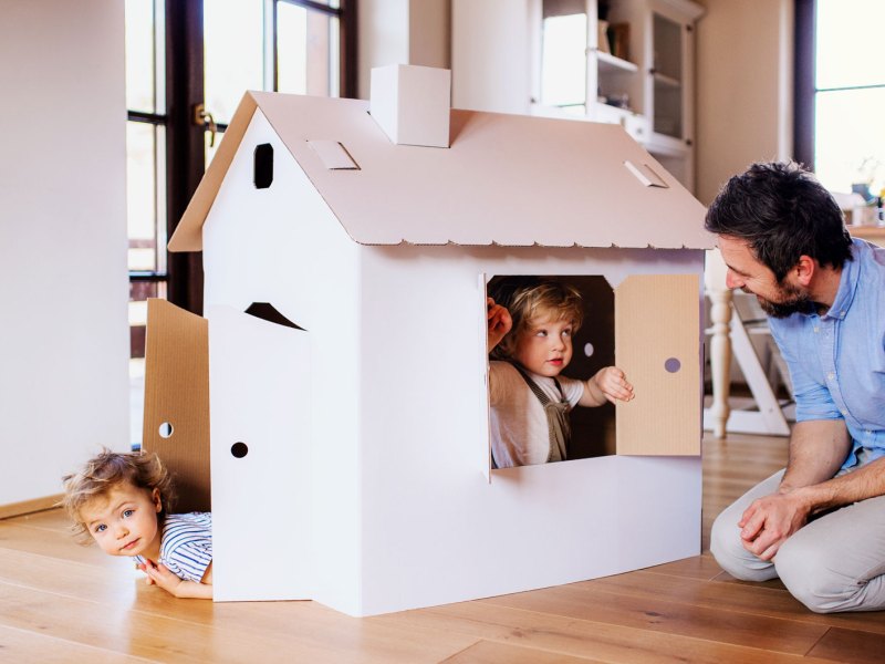 Vater kniet im Wohnzimmer vor dem Pappkartonspielhaus seiner beiden Kleinkinder, die aus Fenster und Tür schauen.