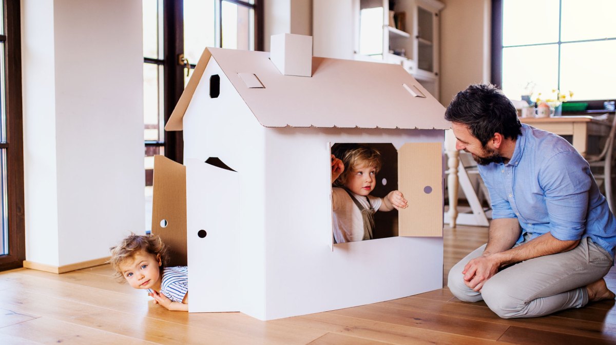 Vater kniet im Wohnzimmer vor dem Pappkartonspielhaus seiner beiden Kleinkinder, die aus Fenster und Tür schauen.