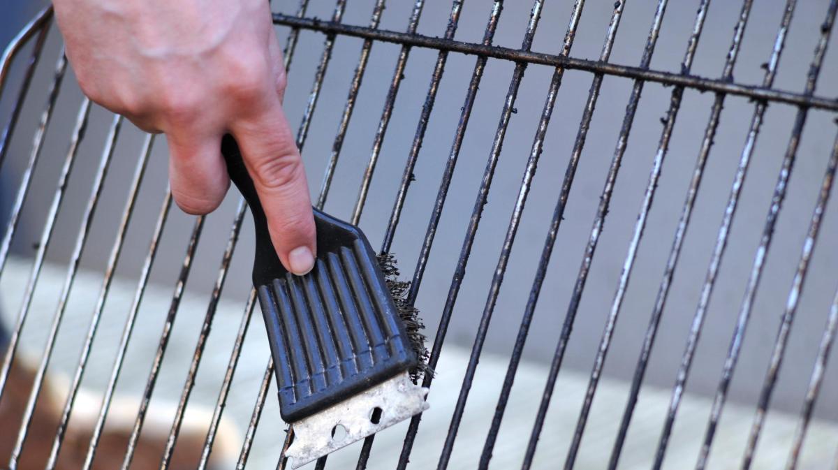 Eine Hand mit Grillbürste reinigt einen stark verschmutzten Grillrost über hellem Boden im Freien bei Tageslicht.