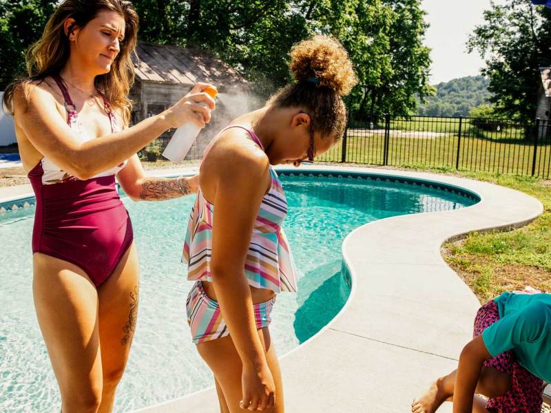 Familie am Pool. Mutter sprüht Tochter mit Sonnenschutz ein. Der kleine Bruder turnt auf einem Stuhl herum.