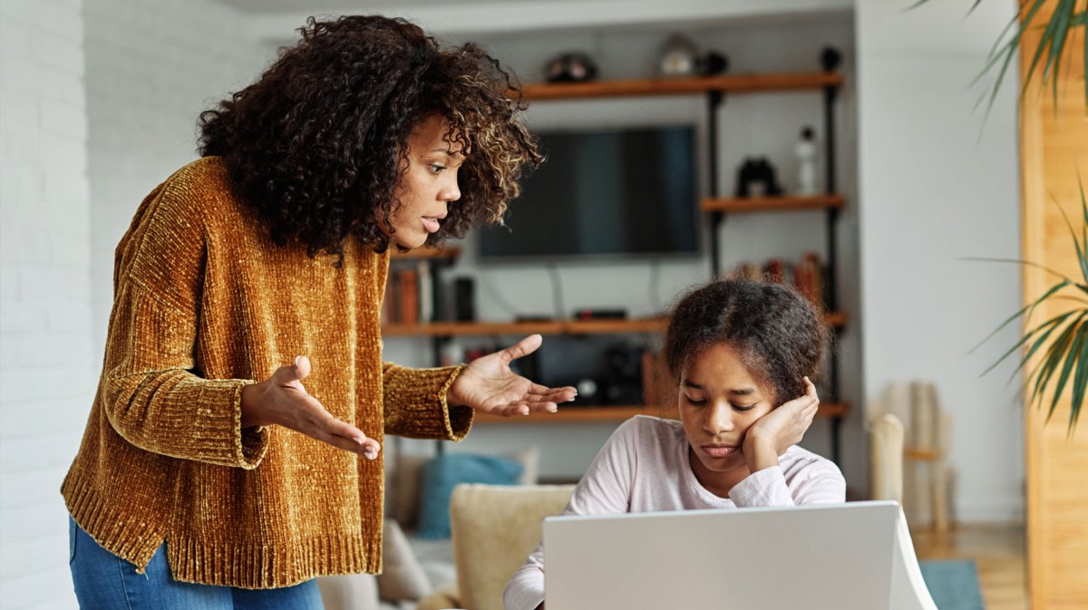 Tochter im Teenageralter sitzt gelangweilt am Laptop, ihre Mutter steht daneben und redet gestikulierend auf sie ein.