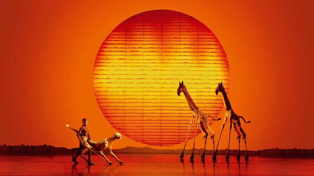 Vor riesiger orange-gelber Sonne ziehen zwei Giraffen-Darsteller anmutig vorbei, ein Mann führt einen Geparden in flacher Savanne.