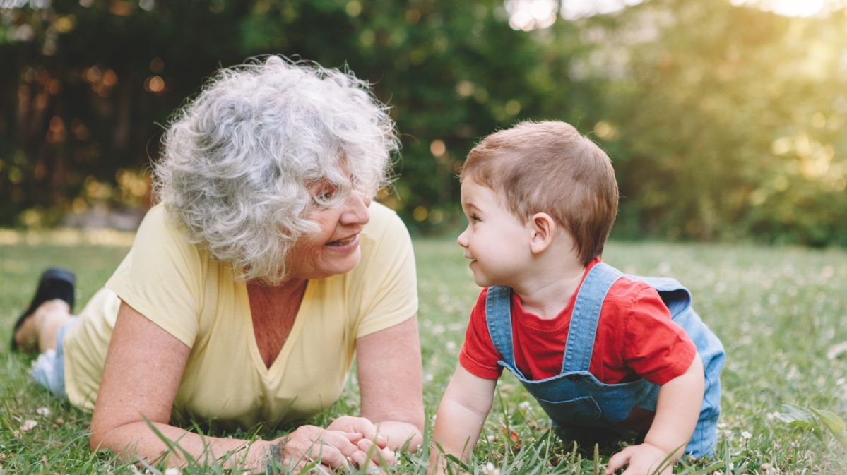 Oma mit grauen Haaren liegt mit ihrem Enkelsohn auf einer Wiese und sie schauen sich an