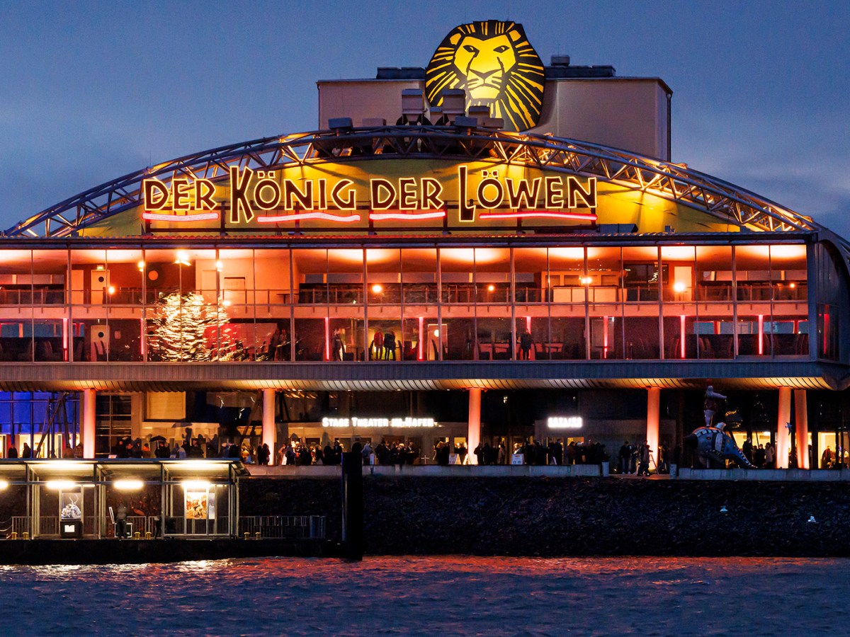 Das hell erleuchtete Stage Theater im Hafen Hamburgs mit „Der König der Löwen“-Schriftzug spiegelt sich im Wasser.