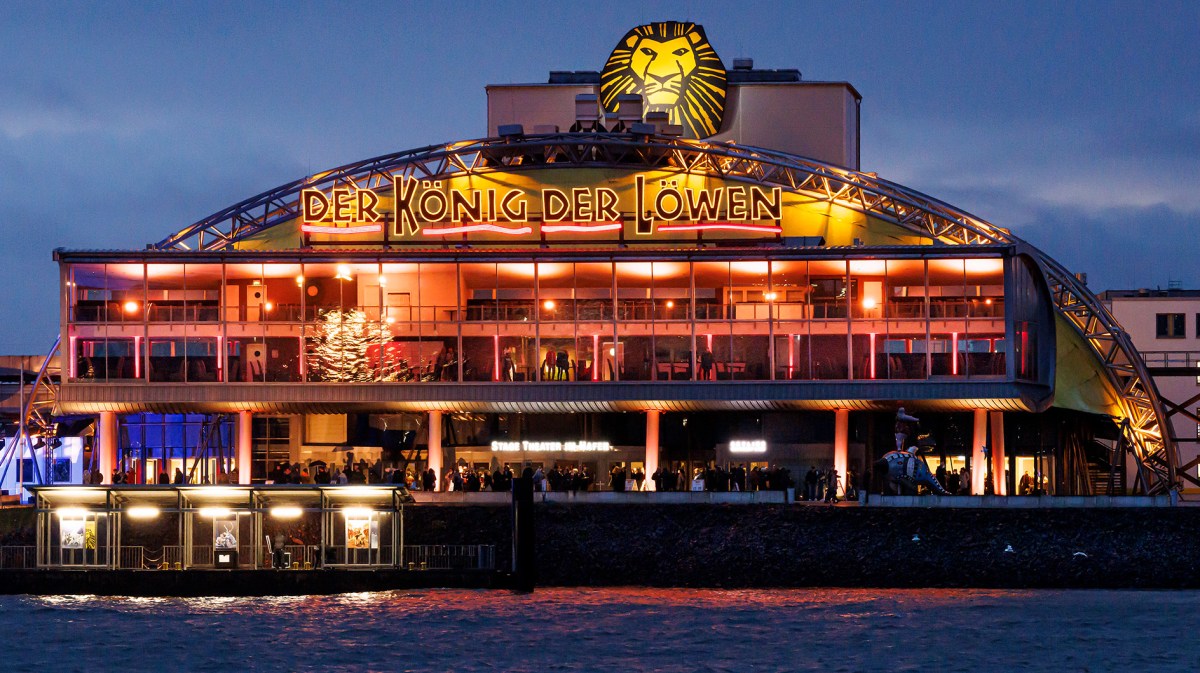 Das hell erleuchtete Stage Theater im Hafen Hamburgs mit „Der König der Löwen“-Schriftzug spiegelt sich im Wasser.