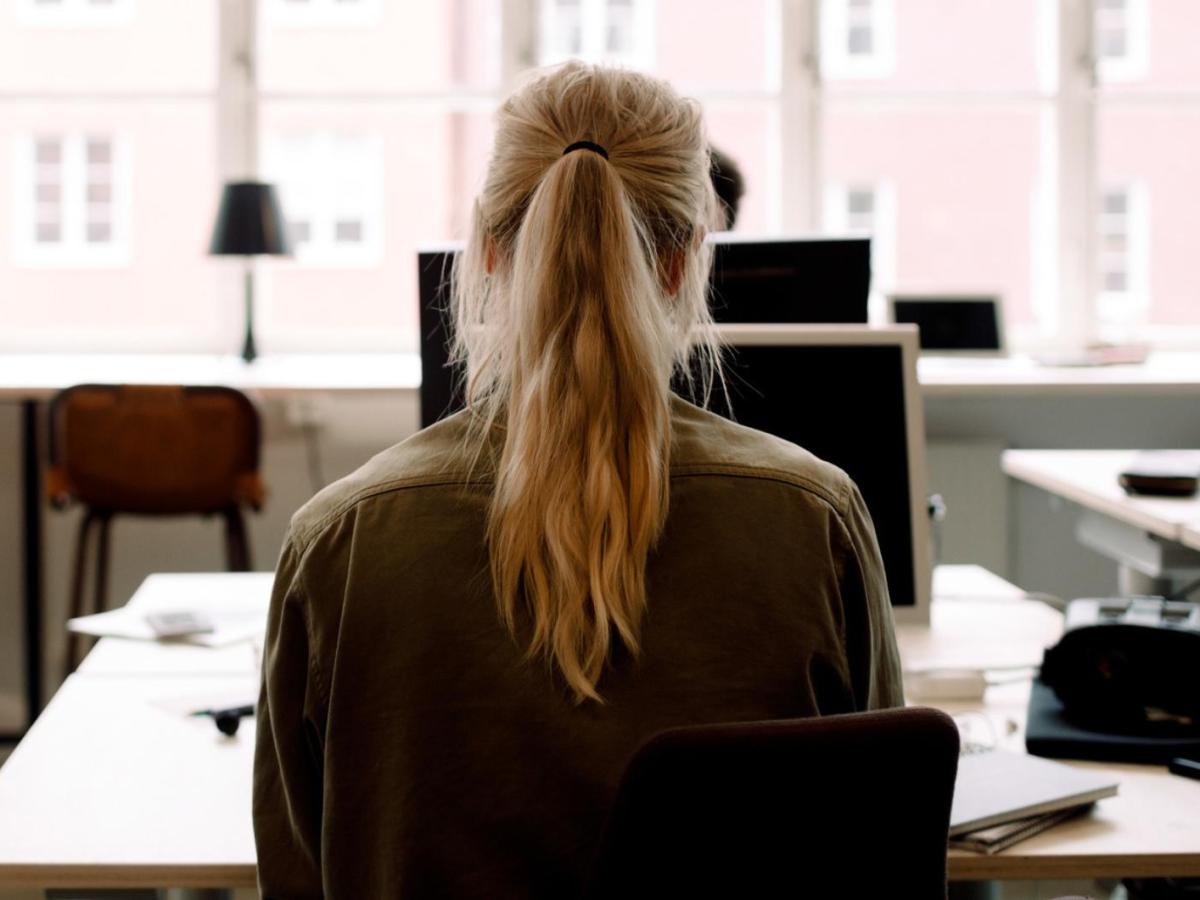 Junge Frau mit einem blonden Pferdeschwanz sitzt an einem Schreibtisch alleine im Büro.