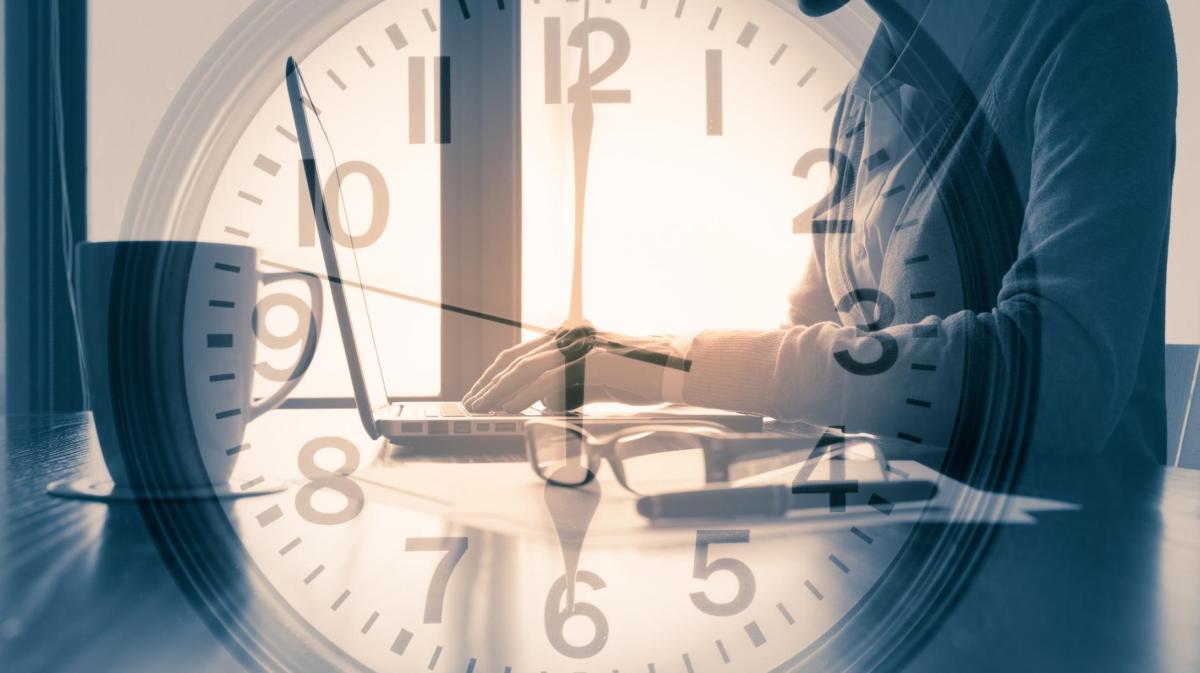 Schattenhafte Frau tippt konzentriert auf Laptop, während große transparente Uhr über dem Bild die Zeit symbolisiert.