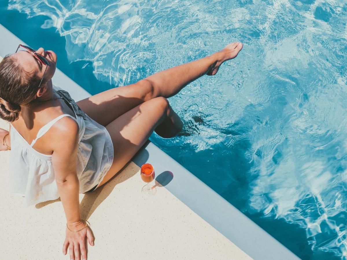 Frau sitzt entspannt am Pool und sonnt sich. Neben ihr steht ein Cocktail.