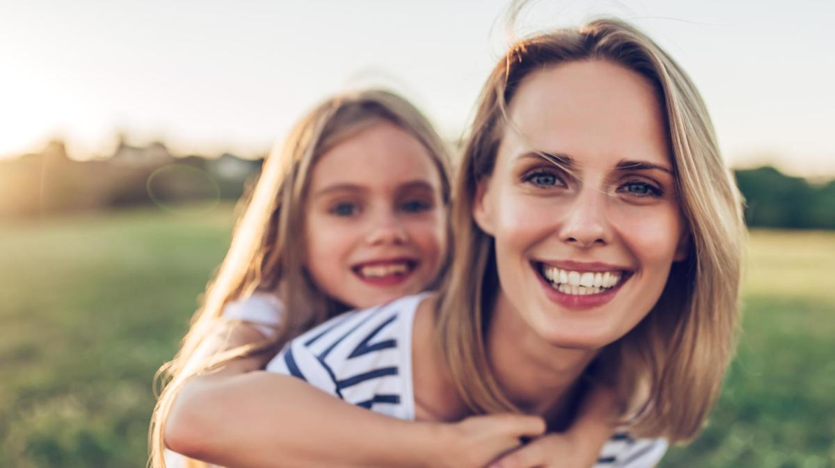 Junge Mutter mit ihrer Tochter. Beide haben blonde lange Haare und spielen draußen zusammen.