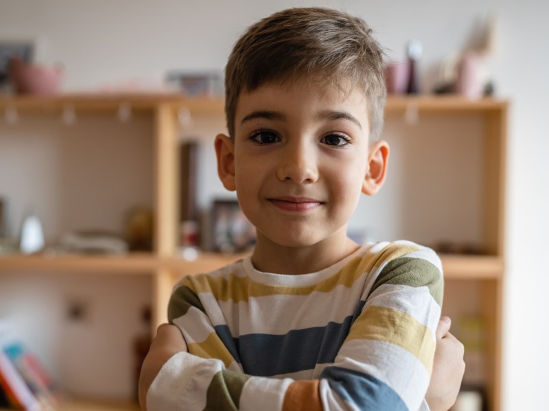 Porträt eines Jungen im Grundschulalter mit verschränkten Armen und aufgesetztem Lächeln.