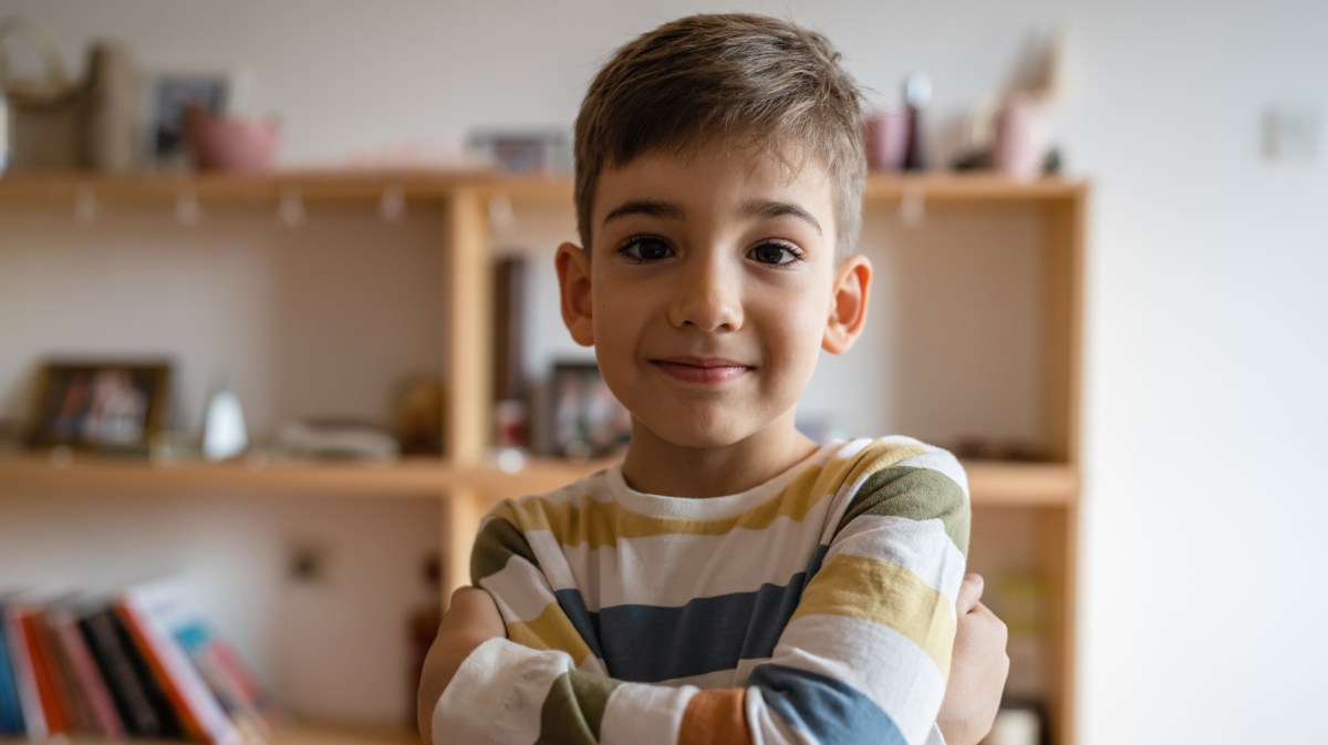Porträt eines Jungen im Grundschulalter mit verschränkten Armen und aufgesetztem Lächeln.