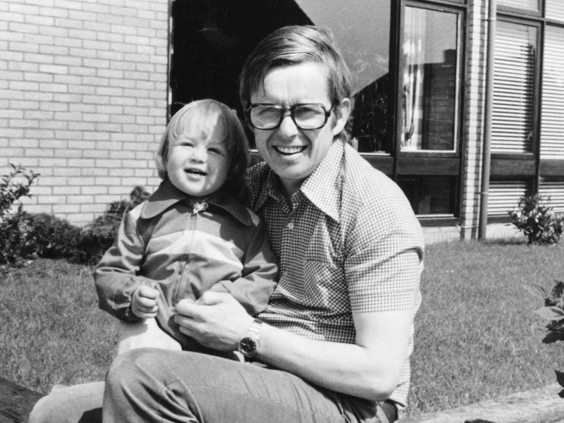 Vintage-Foto von 1978: Ein Vater sitzt mit seinem ca. 2-jährigen Sohn vor einem Einfamilienhaus.