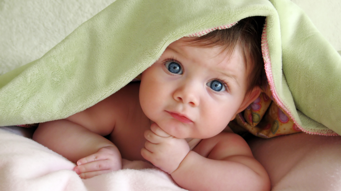 Baby mit blauen Augen liegt auf dem Boden und schaut unter der Decke hervor, direkt in die Kamera.