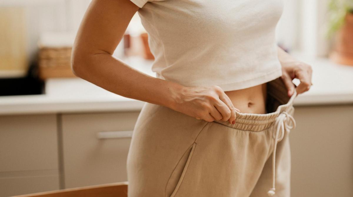 Frau hat eine zu weite Jogginghose mit Gummizug an und zeigt mit den Händen, wie viel Platz die Hose oben am Bund hat.