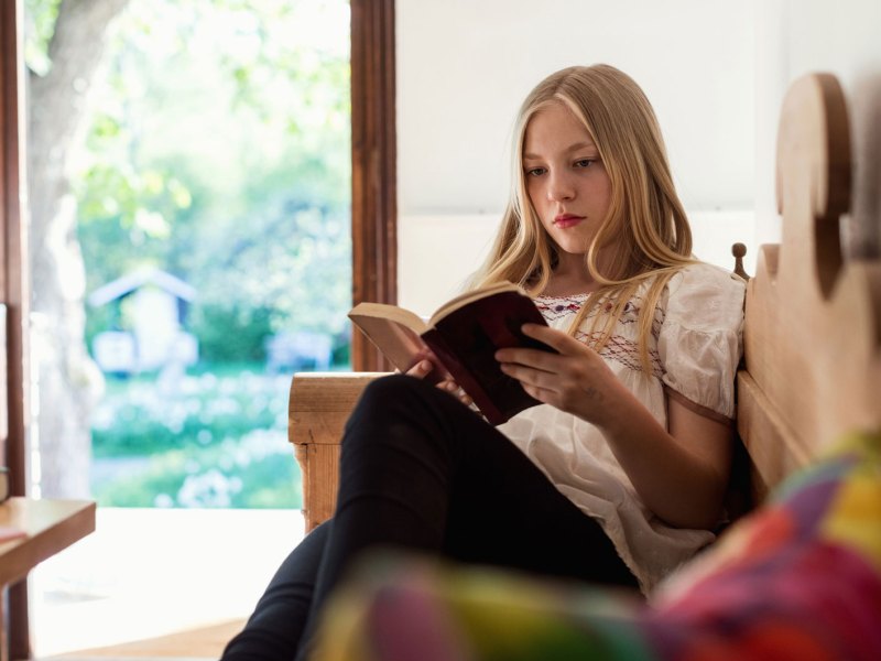 Teenagerin liest ein Buch, während sie drinnen auf einer Bank sitzt.
