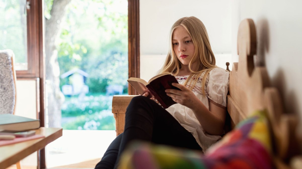Teenagerin liest ein Buch, während sie drinnen auf einer Bank sitzt.
