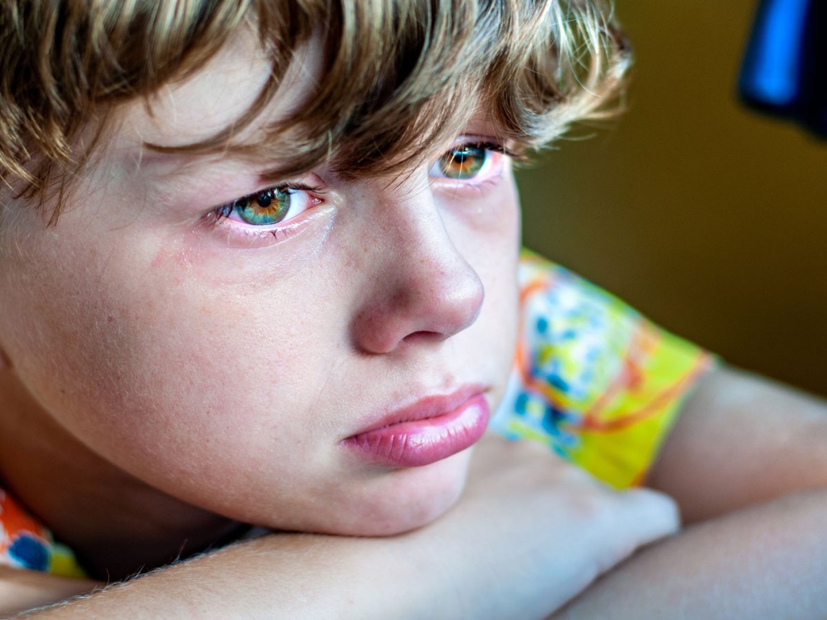 Profil eines traurigen Jungen, der den Kopf auf den auf einem Tisch liegenden verschränkten Armen abgelegt hat.