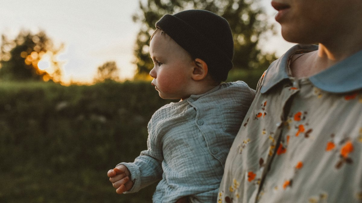 Baby wird bei Sonnenuntergang draußen getragen, trägt eine stylische Mütze und ein Hemd.