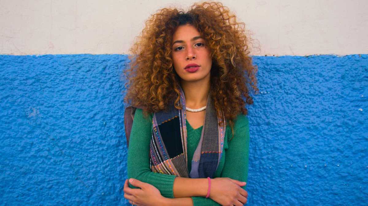 Junge Frau mit Locken steht vor einer blau-weißen Wand und schaut in die Kamera.