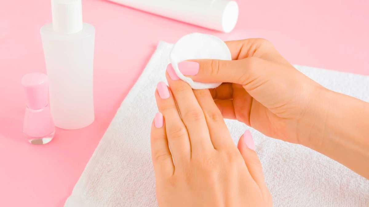 Junge Frau entfernt rosa Nagellack mit einem weißen Wattepad auf einem Handtuch auf einem hellrosa Tischhintergrund. Nahaufnahme.