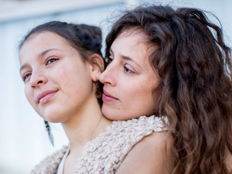 Nahaufnahme von Mutter und Tochter. Die Mutter umarmt ihre Tochter von hinten und legt ihren Kopf auf die Schulter der Tochter.