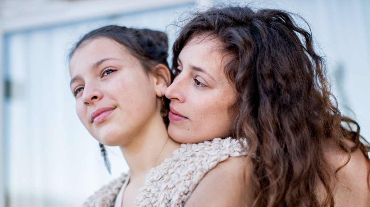 Nahaufnahme von Mutter und Tochter. Die Mutter umarmt ihre Tochter von hinten und legt ihren Kopf auf die Schulter der Tochter.