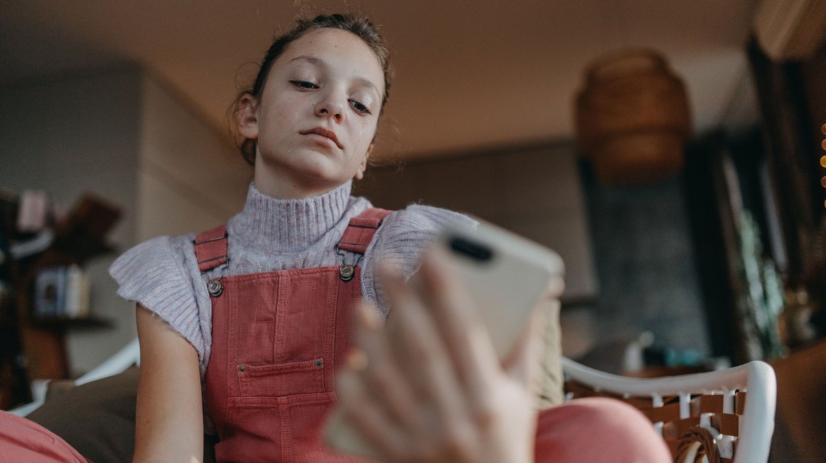 Mädchen im Teenageralter sitzt auf dem Sofa und schaut genervt auf ihr Smartphone.