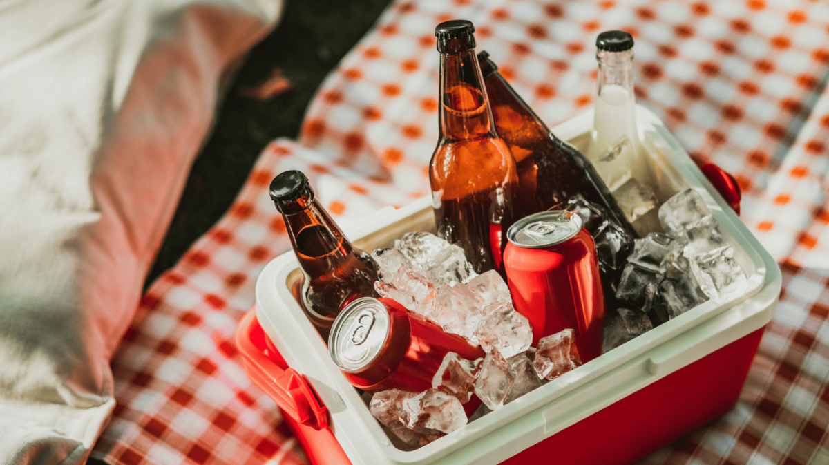 Kühlbox mit Eiswürfel, Bierflaschen und Coladosen auf Picknickdecke.