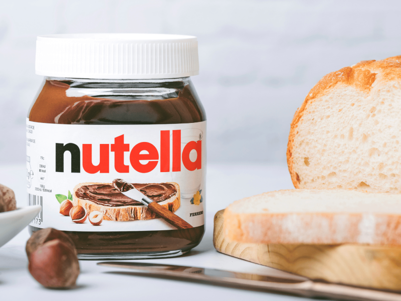 Nutella-Glas auf Tisch, neben Weißbrot und Haselnüssen.