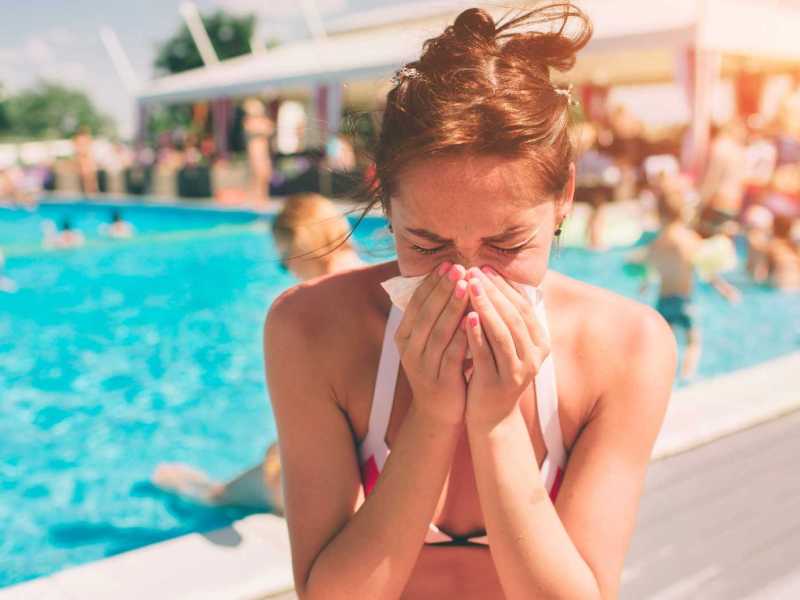 Frau sitzt krank am Pool im Bikini und schnäuzt sich die Nase