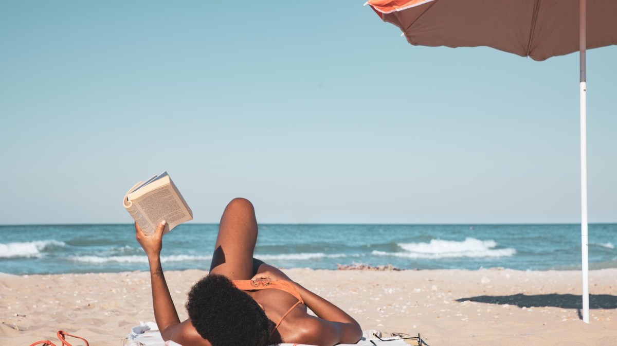 Frau liegt am Strand unter Sonnenschirm und liest einen Kurzroman