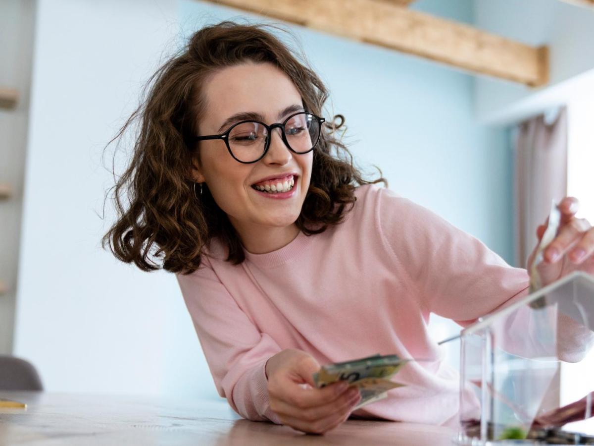 Frau mit Brille und rosa Shirt wirft Geld in eine Spardose und lacht.