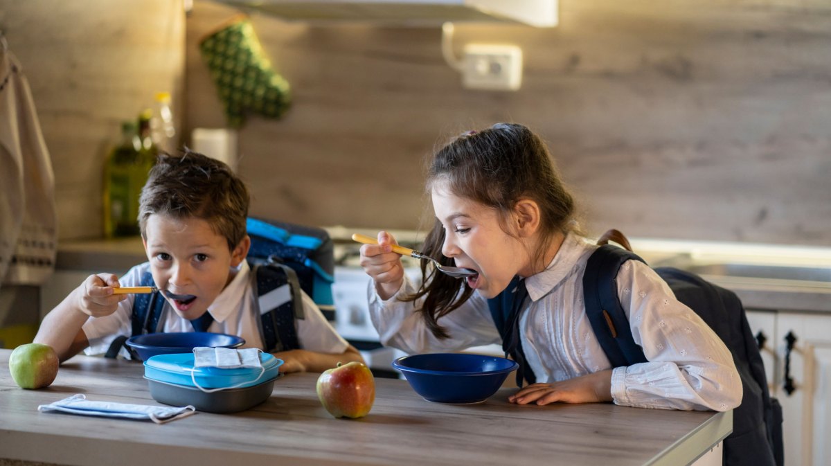 Zwei Schulkinder in Schuluniform sitzen am Tisch und essen hektisch ein Müsli.
