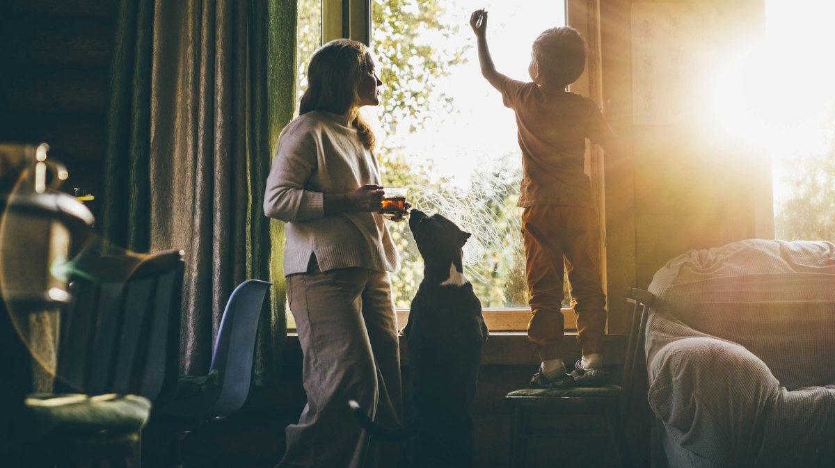 Mutter, Kind und Hund stehen in Pyjamas morgens am Fenster, die Sonne geht gerade auf und schauen nach draußen.