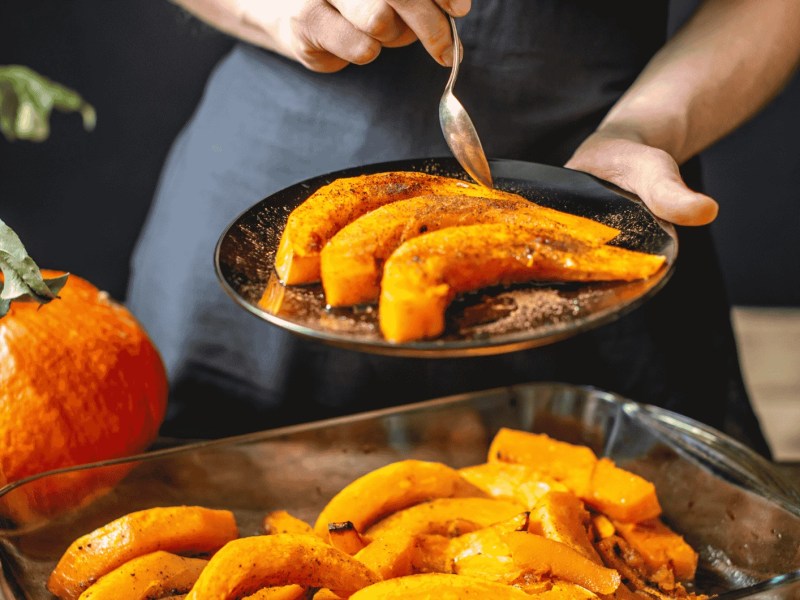 Köchin hält Scheiben von gebackenem orange Hokkaido-Kürbis mit Honig und Zimt. Herbstliches Essen in einer gemütlichen Küche.