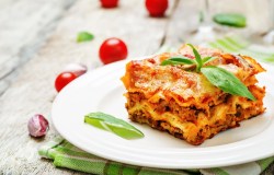 eine Portion Lasagne auf einem weißen Teller, Tomaten und Basilikum auf dem Tisch