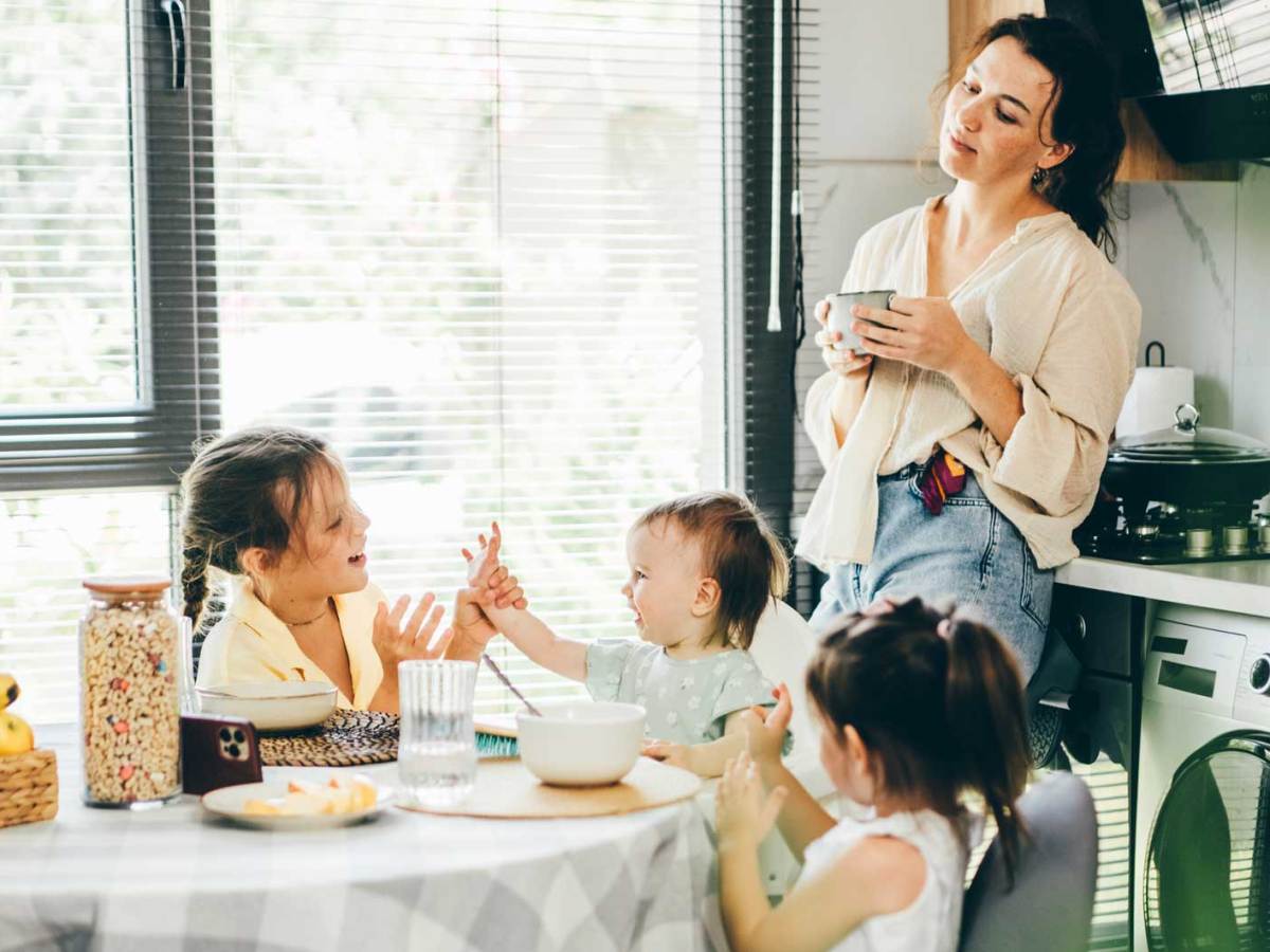 Mutter steht in der Küche und beobachtet ihre drei Kinder, die am Frühstückstisch sitzen und miteinander agieren.