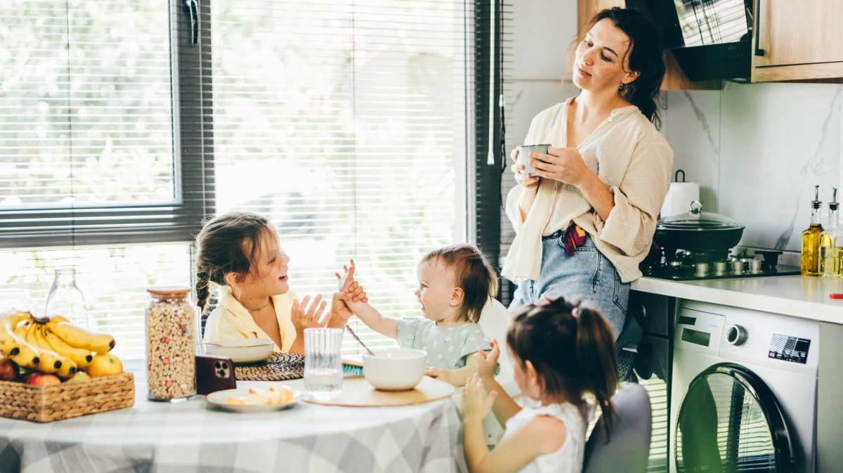 Mutter steht in der Küche und beobachtet ihre drei Kinder, die am Frühstückstisch sitzen und miteinander agieren.