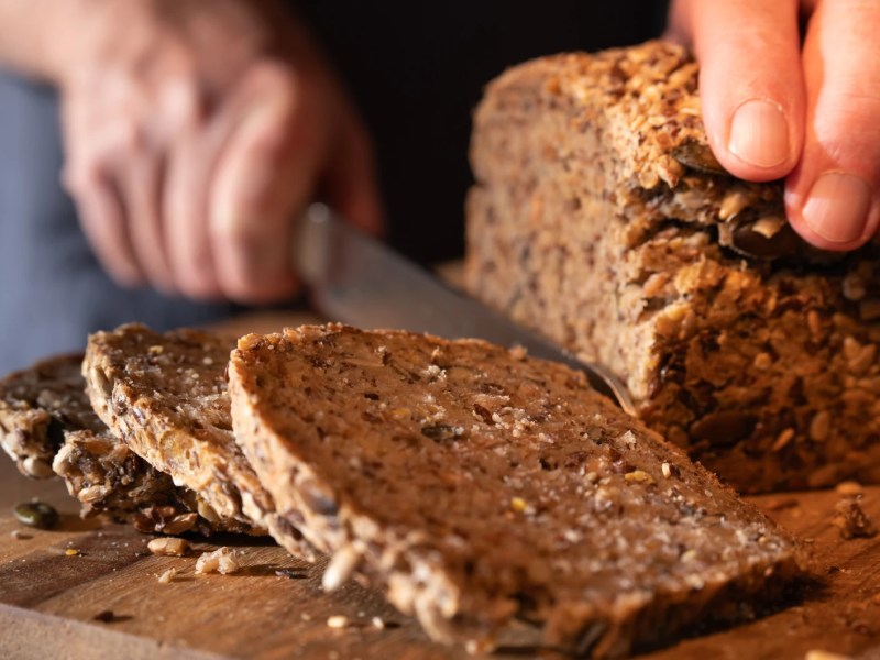 Nahaufnahme von Männerhänden, die Vollkornbrot auf einem Brett schneiden.