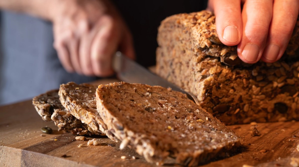 Nahaufnahme von Männerhänden, die Vollkornbrot auf einem Brett schneiden.