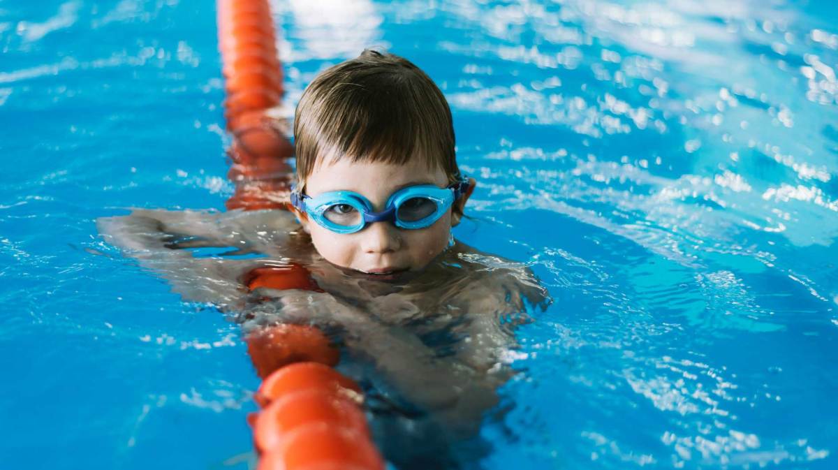 Junge, ca 6 Jahre alt, guckt mit Schwimmbrille aus dem Wasser eines Schwimmbads.
