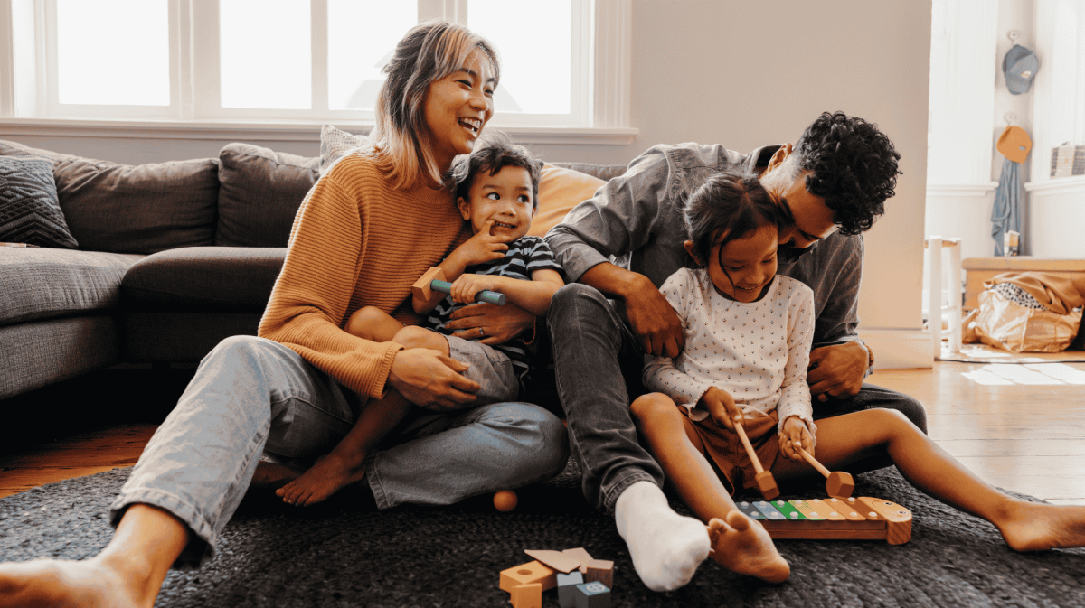 Familie mit zwei Kindern sitzt auf Boden vor Sofa und lacht.
