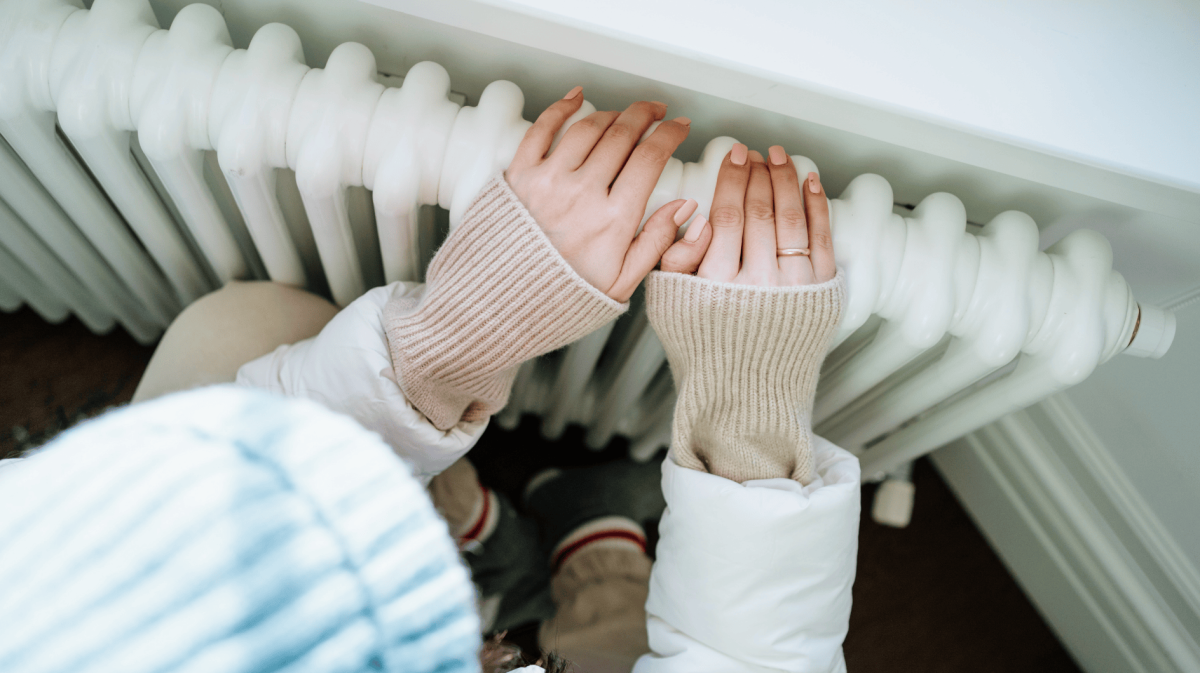 Frau sitzt warm angezogen vor der Heizung und hält ihre Hände auf die Heizung.