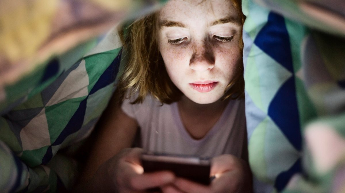 Mädchen im Teenageralter liegt im Bett unter der Decke und schaut auf ihr Handy.